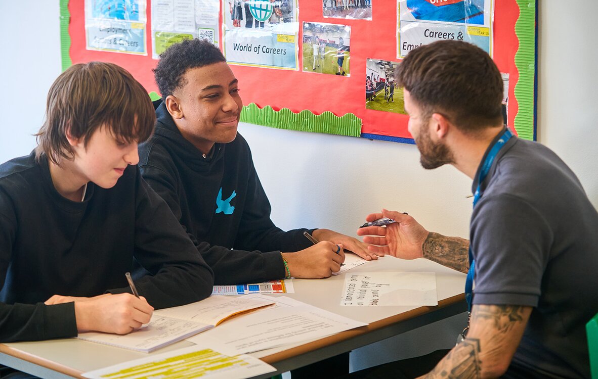 two pupils sat at a desk opposite an adult