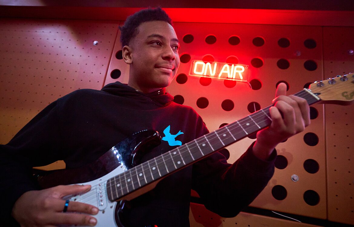 pupil playing guitar in front of on air sign
