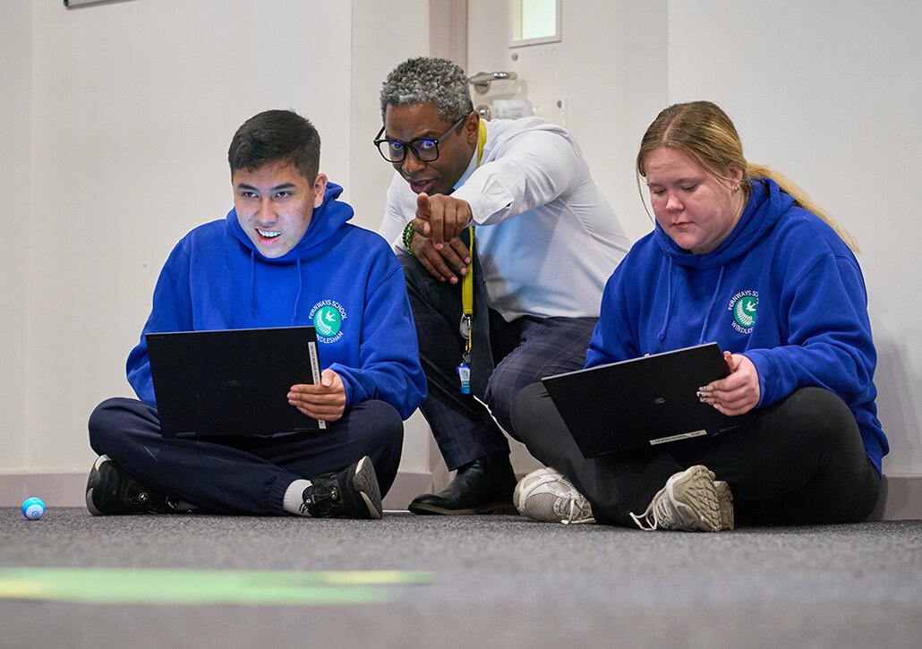 pupils with laptops take instruction from a teacher