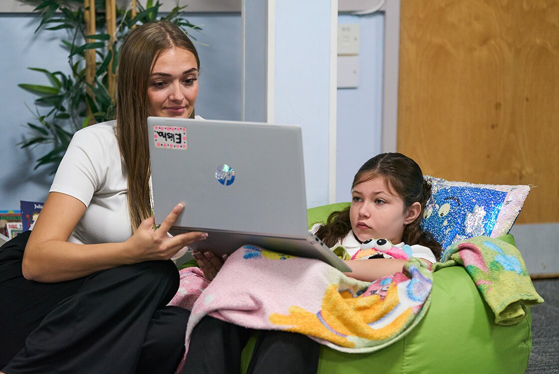 child works on laptop whilst sat on a bean bag supported by a member of staff