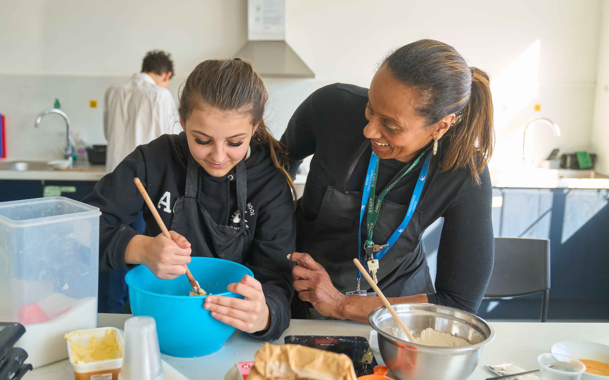 girl stirs ingredients in mixing bowl watched by adult