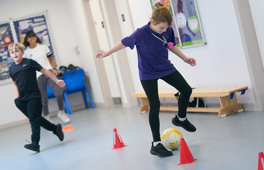 Pupils dribbling a football around cones indoors