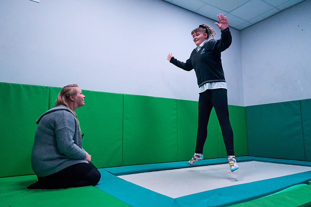 A pupil bounces on a trampoline indoors while a teacher kneels watching
