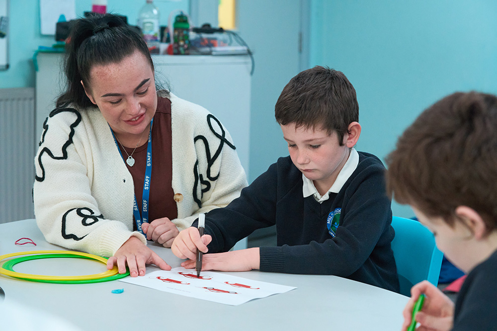 Pupil writes with pen on paper with a teaching assistant sat at a table