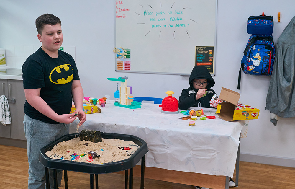 pupil standing plays with sand in a table top tray whilst another plays with playdoh