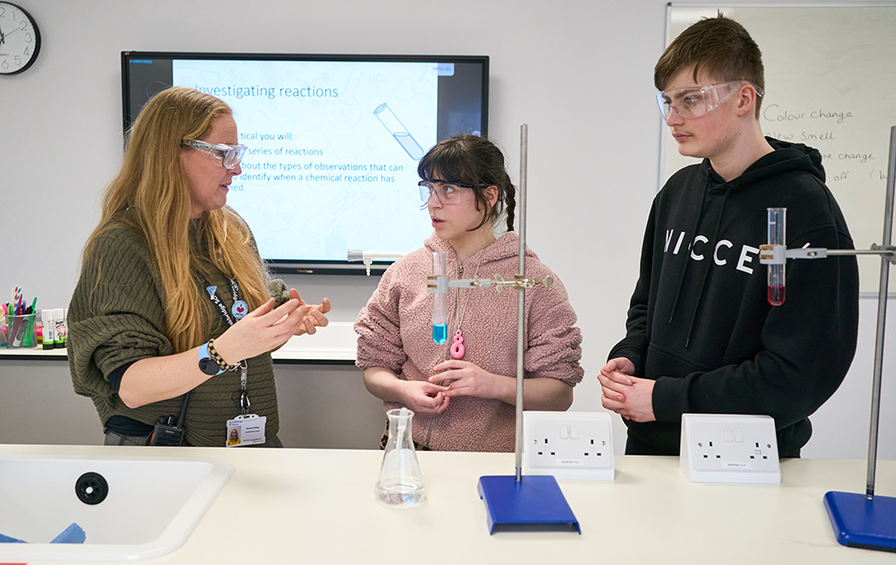 two pupils talk wit ha teacher behind a science bench and equipment