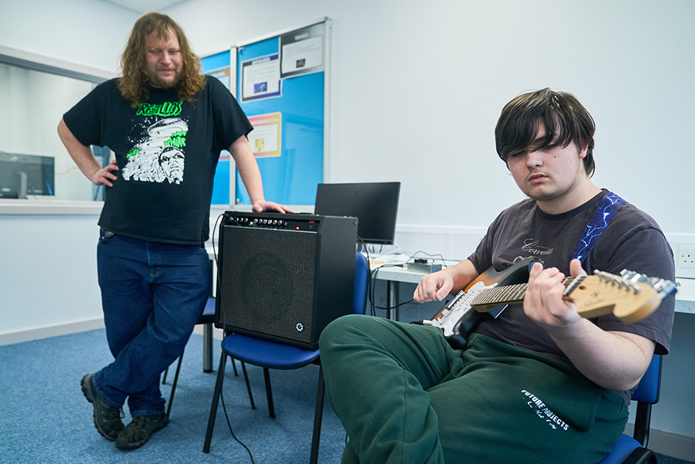 boy plays electric guitar watched by adult
