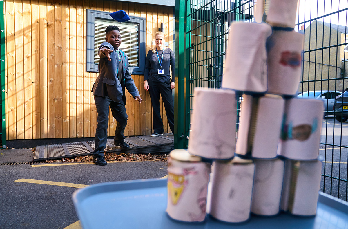 child throws bean bag at stack of tins watched by an adult